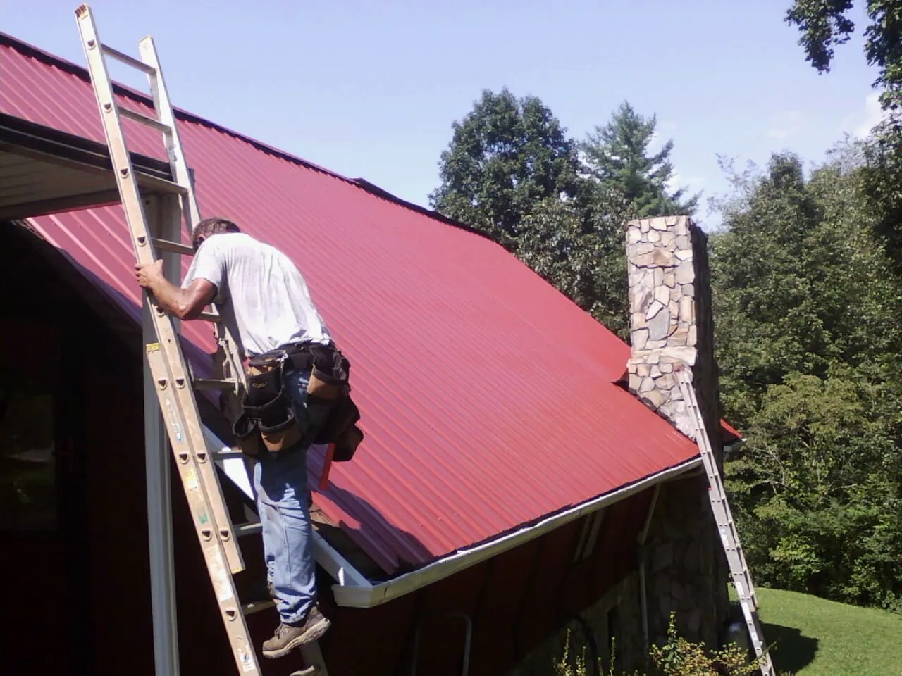 Roofer on a ladder beside a red metal roof and stone chimney — Soffit & Fascia Repair by Roofing Brothers in Floyd